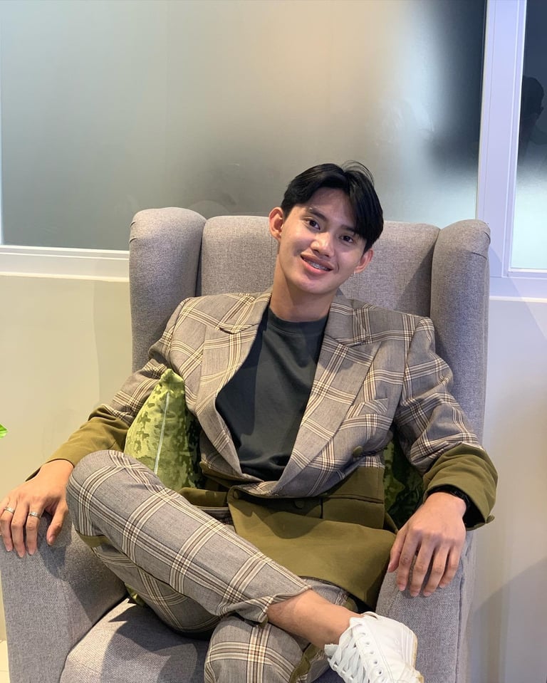 Young man in beige plaid blazer and pants sitting in gray armchair, smiling at camera indoors