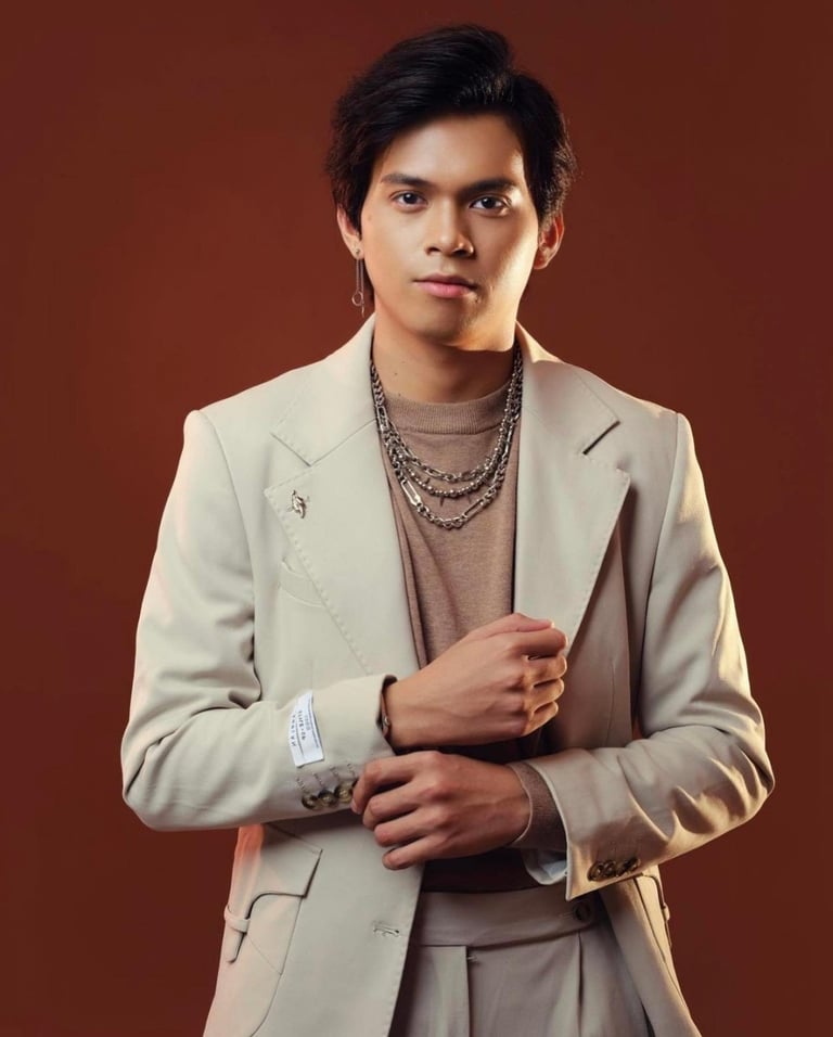 Young man wearing a beige blazer and layered chain necklaces poses against a brown background