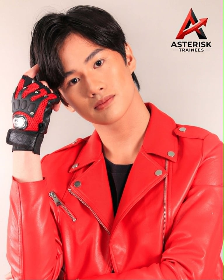 Person wearing bright red leather jacket and red-black gloves, touching face, with Asterisk Trainees logo visible in upper right