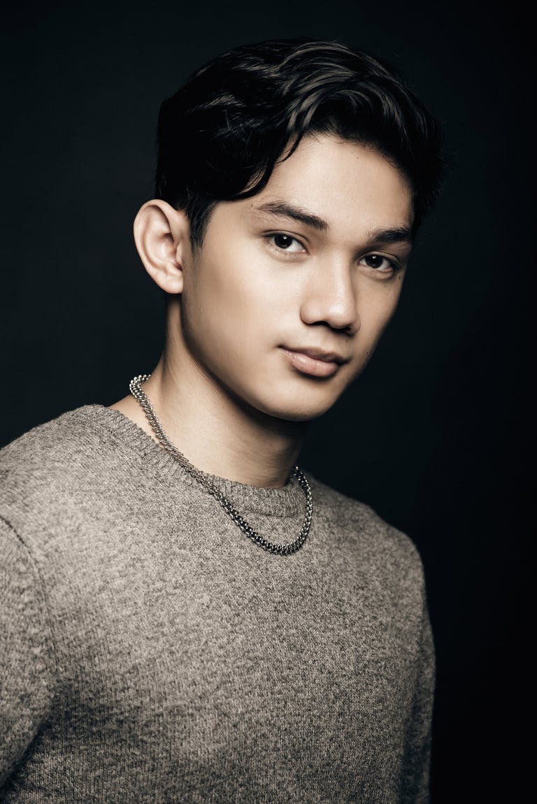 Young man with dark hair wearing a gray sweater and chain necklace against a dark background