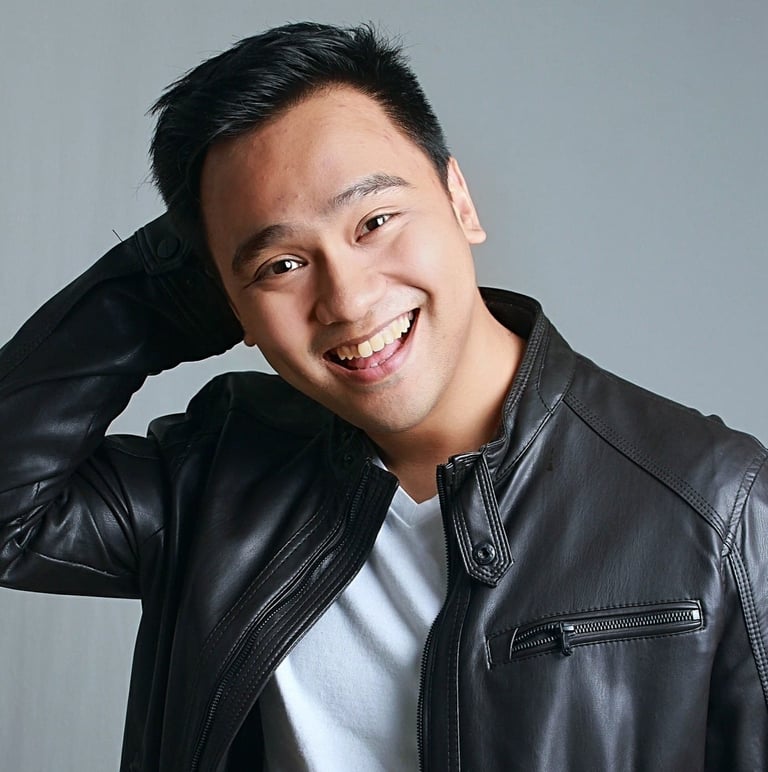 Smiling man wearing black leather jacket and white shirt, posing with hand near head against gray background