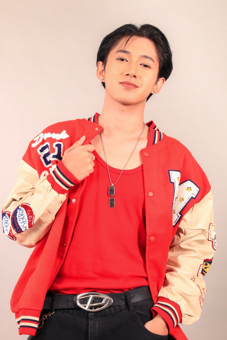 Young Asian man wearing red and cream varsity jacket with patches, red shirt, and dog tag necklace, posing against beige background