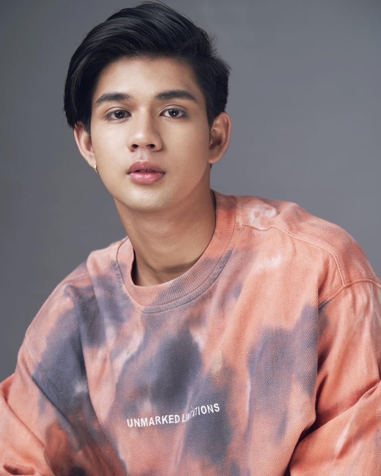 Young man with dark hair wearing a tie-dye pink and purple sweatshirt against a gray background