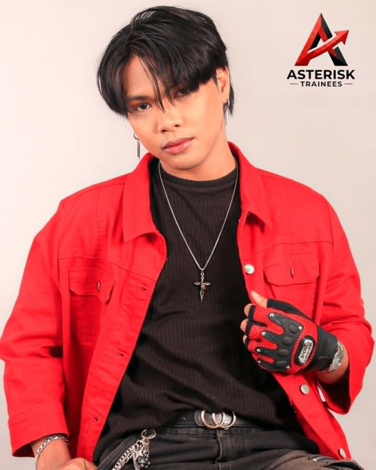 Person wearing red jacket and black shirt with cross necklace and red gloves against beige background with Asterisk Trainees logo
