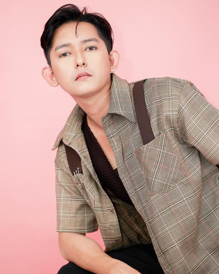 Man wearing a beige plaid shirt over a black shirt poses against a pink background