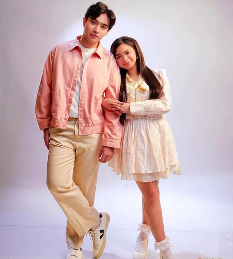 Young man in pink jacket and beige pants standing with a young woman in cream dress against a white background