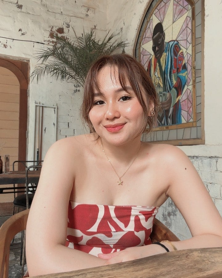 Young woman smiling at camera indoors wearing red and white strapless top with potted plant and stained glass window behind her