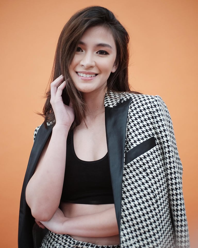 Young woman smiling at camera wearing black crop top and houndstooth blazer against orange background