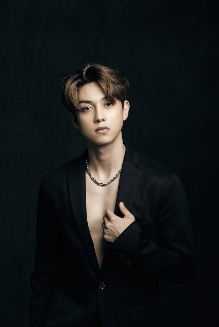 Young man with light brown hair wearing a black blazer and chain necklace against a dark background in a portrait studio setting