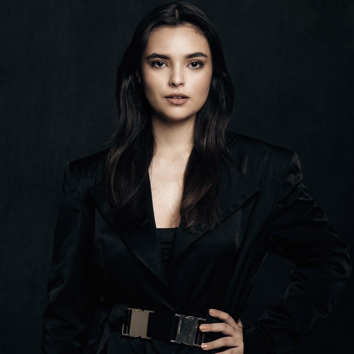 Woman with long dark hair wearing a black blazer and belt against a dark background, looking directly at camera
