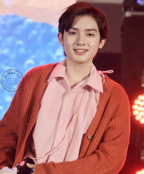 Young man smiling at camera wearing pink button-up shirt and red cardigan on stage with blue background