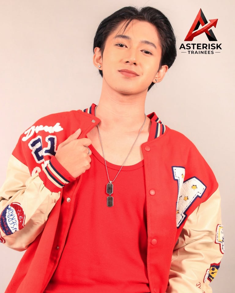 Young man wearing red and cream varsity jacket with Asterisk Trainees logo, posing against beige background