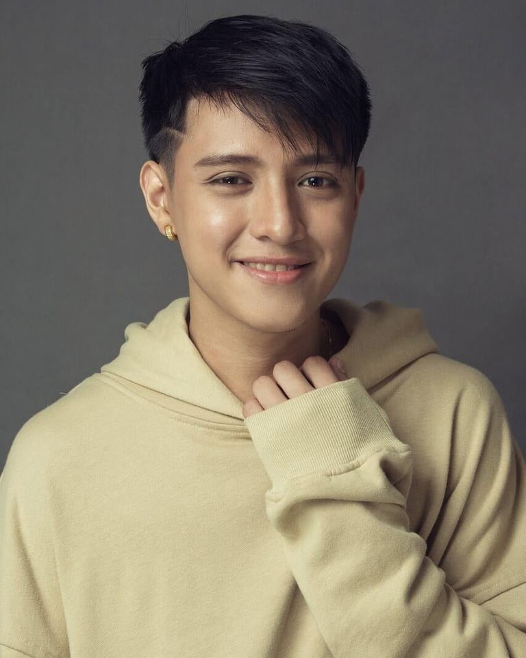 Young person wearing a cream-colored hoodie, smiling at the camera against a gray background. They have short dark hair and gold ear piercings.