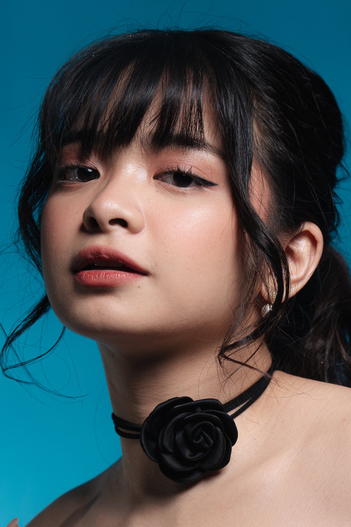 Portrait of a young woman with black bangs and long dark hair wearing a black rose pendant necklace against a bright teal background