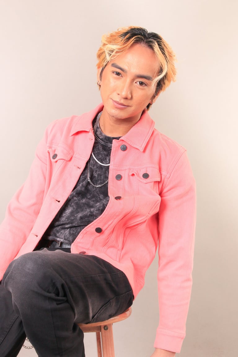 Young person with blonde and black hair wearing a pink jacket and dark clothing, sitting on a stool against a neutral background
