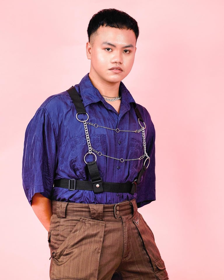 Young person wearing a blue button-up shirt with chain harness and brown pants against a pink background