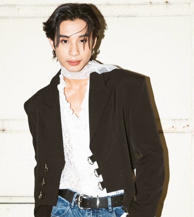 Person wearing black blazer over white lace shirt with blue jeans, standing against white wall
