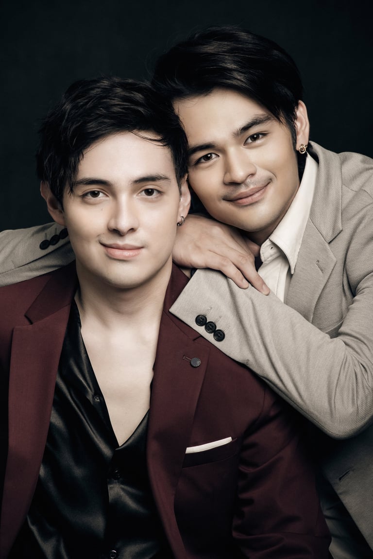 Two young men posing together against a dark background, one wearing a burgundy blazer and the other in a beige blazer, smiling at the camera.