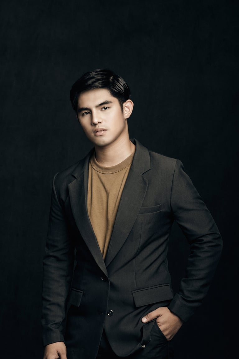 Young man in black blazer and tan shirt posing against dark background, hands in pockets, looking at camera