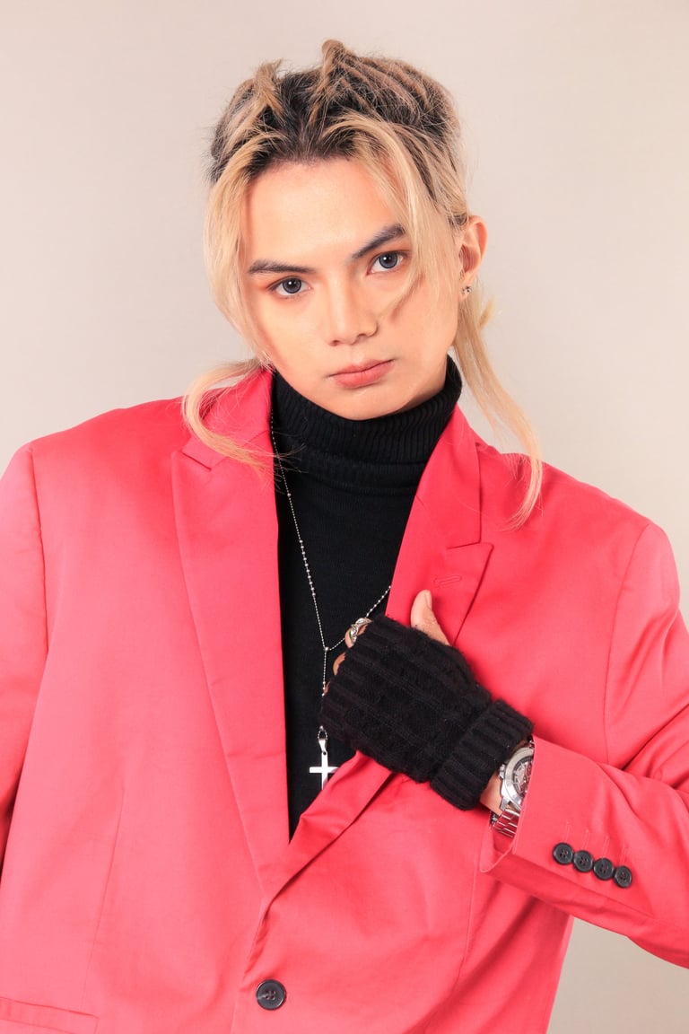 Young person with blonde and dark hair wearing a bright pink blazer and black turtleneck, posing against a neutral background