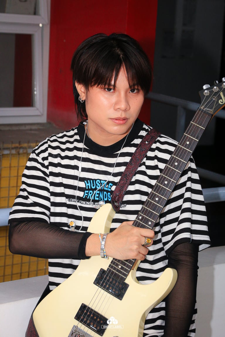 Young person with dark hair wearing striped black and white shirt holding a cream-colored electric guitar indoors