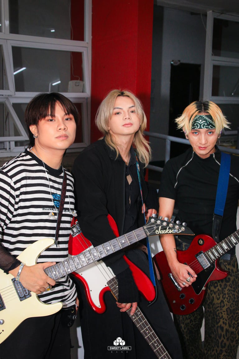 Three musicians with guitars posing in front of a red wall in an indoor venue setting