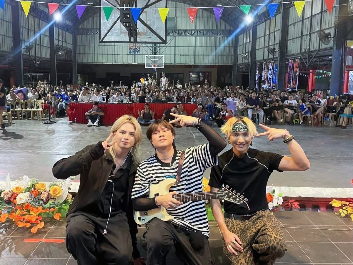 Three people posing in front of a large crowd gathered in an indoor arena with colorful bunting and decorations overhead.