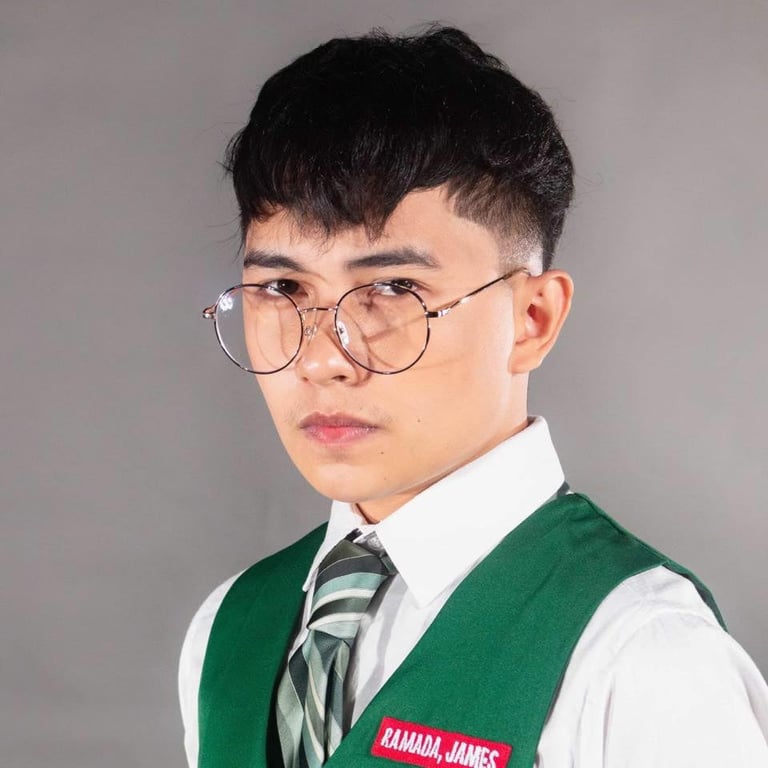 Young man wearing glasses, white dress shirt, green vest with name tag, and striped tie against neutral background
