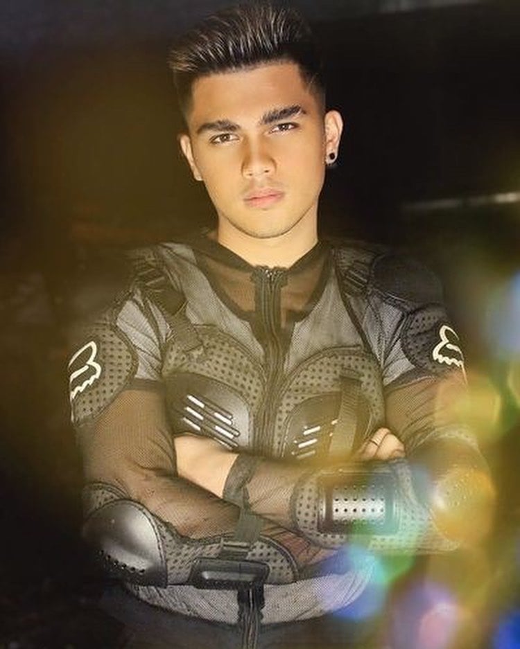Young man with dark spiky hair wearing a black mesh jacket with arms crossed, looking directly at camera with glowing light effects