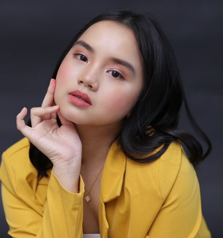Woman in yellow shirt with hand near face, dark hair, against gray background
