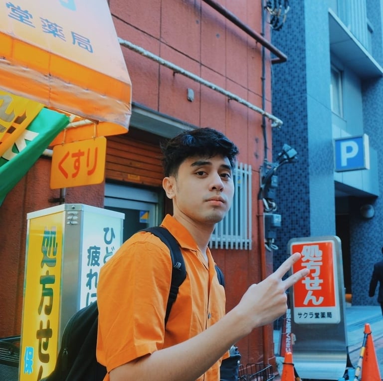 Young man in orange shirt with backpack standing on urban street with Japanese signage and storefronts