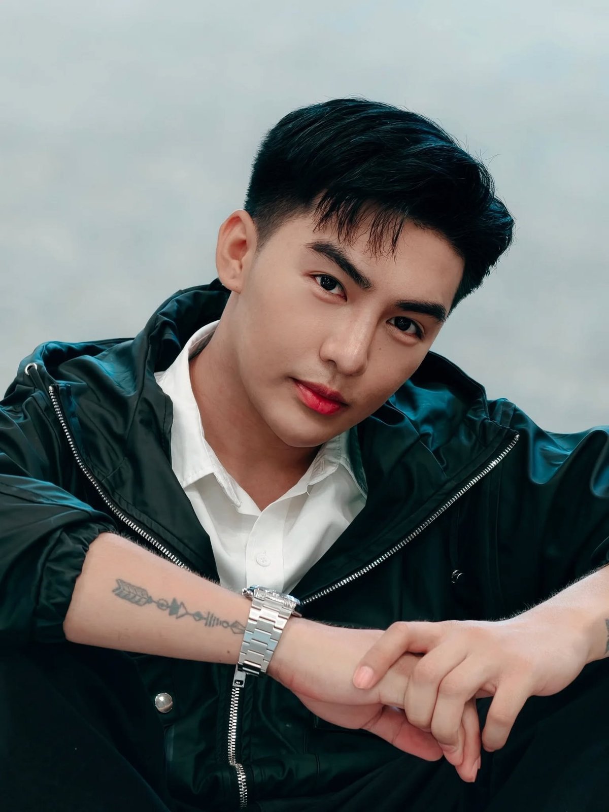 Young man with dark hair wearing a black jacket and white shirt, displaying a tattoo on his arm with a watch