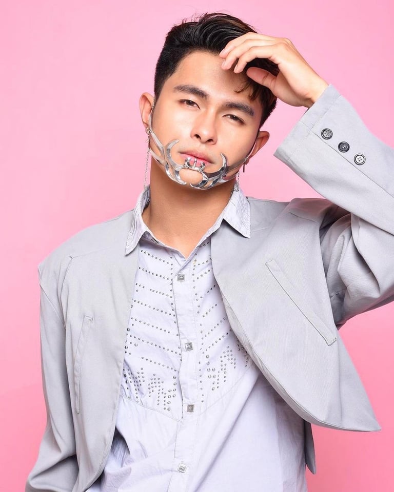 Young man in light gray blazer and patterned shirt with plastic wrap on face against pink background, hand touching head