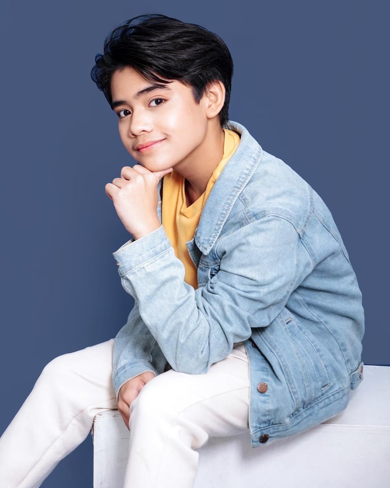 Young person with black hair wearing a light blue denim jacket and yellow shirt, sitting against a blue background with hand near face.