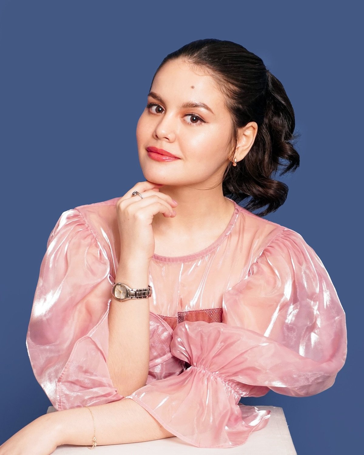 Woman with dark wavy hair wearing a pink velvet top against a blue background, posed with hand near shoulder