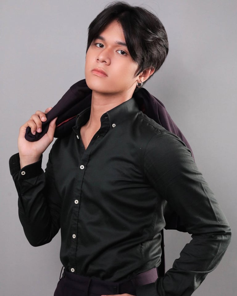 Young man with dark hair wearing a black button-up shirt, posing against a gray background with hand on shoulder