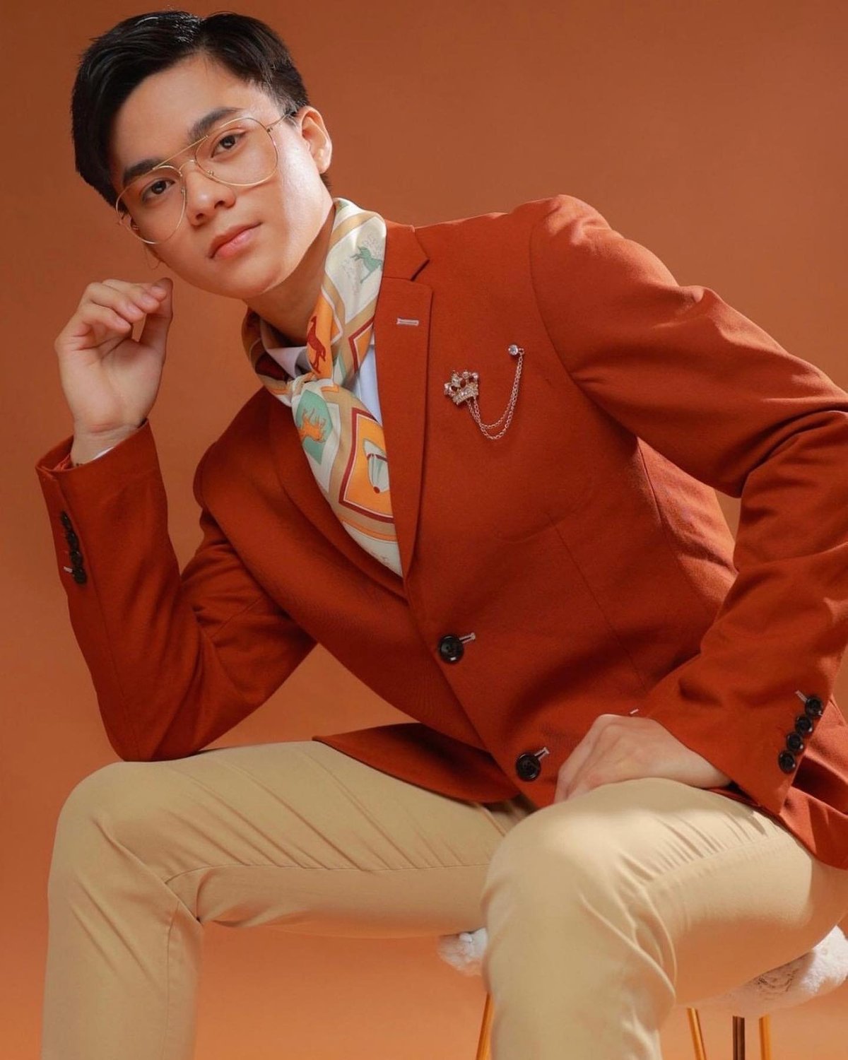 Person wearing clear glasses and a rust-red blazer over a colorful patterned shirt, posing against an orange background