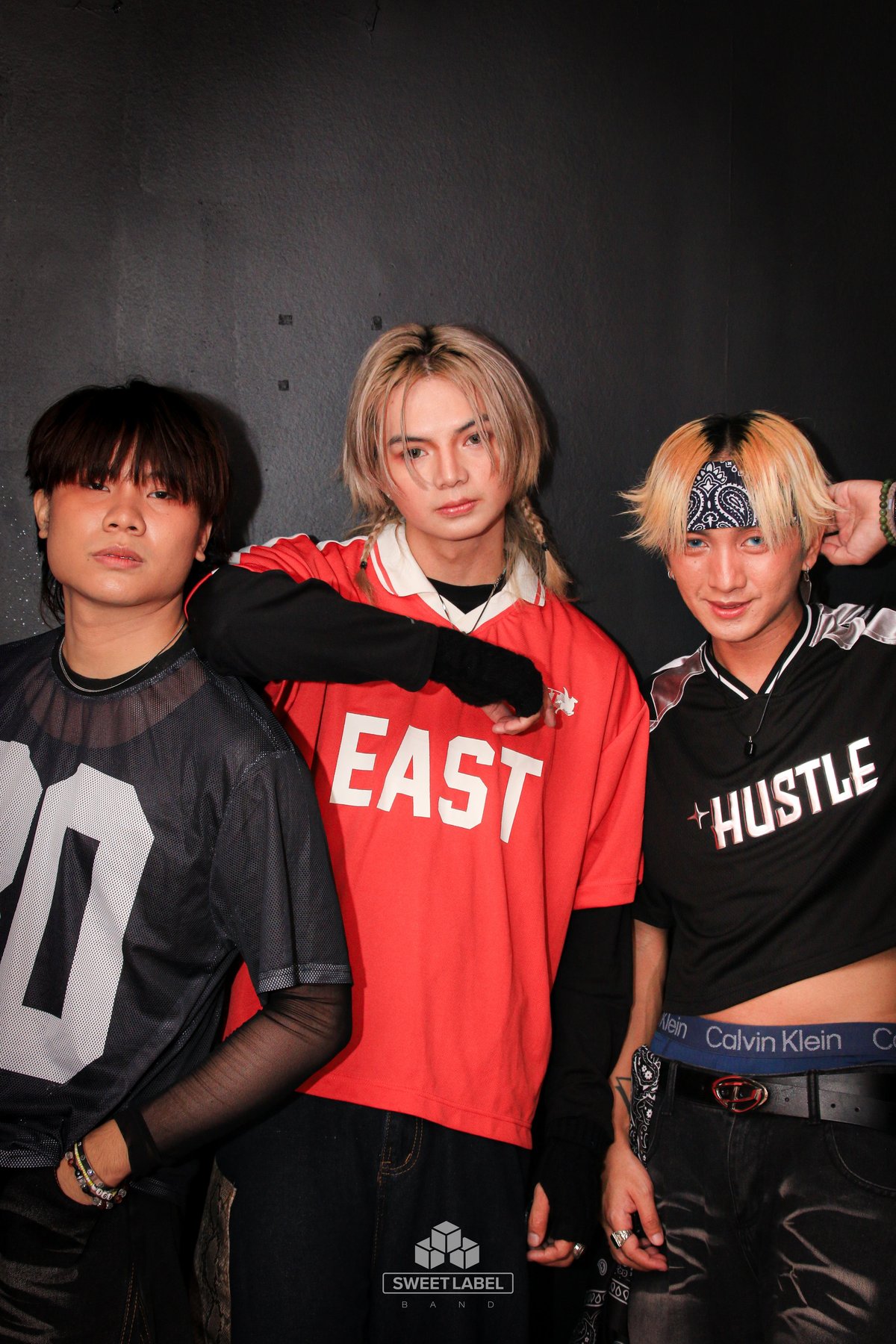 Three people posing for a photo against a dark gray backdrop. From left to right: person in black graphic shirt, person in red EAST hoodie, and person in black HUSTLE shirt with blonde and black hair.