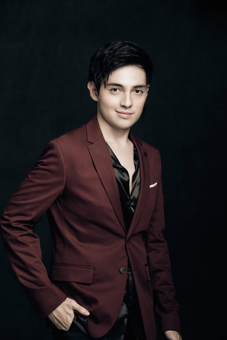 Young man in burgundy blazer and black shirt standing against dark background with hand in pocket