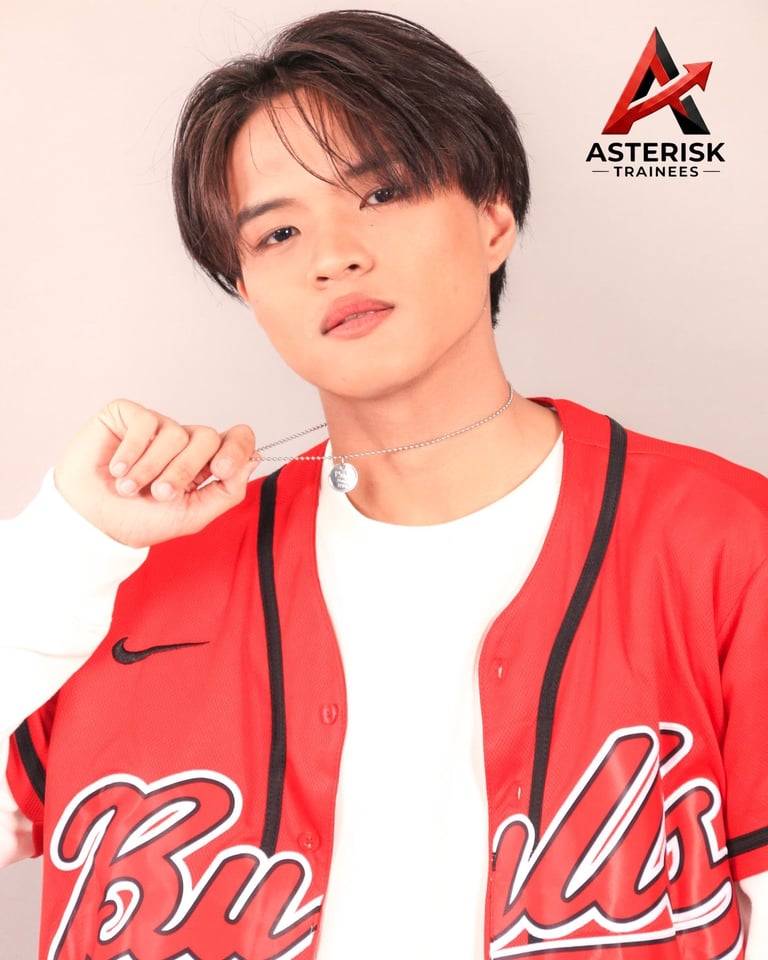 Young person with dark hair and colorful face makeup wearing a red Nike baseball jacket against a cream background with Asterisk Trainees logo