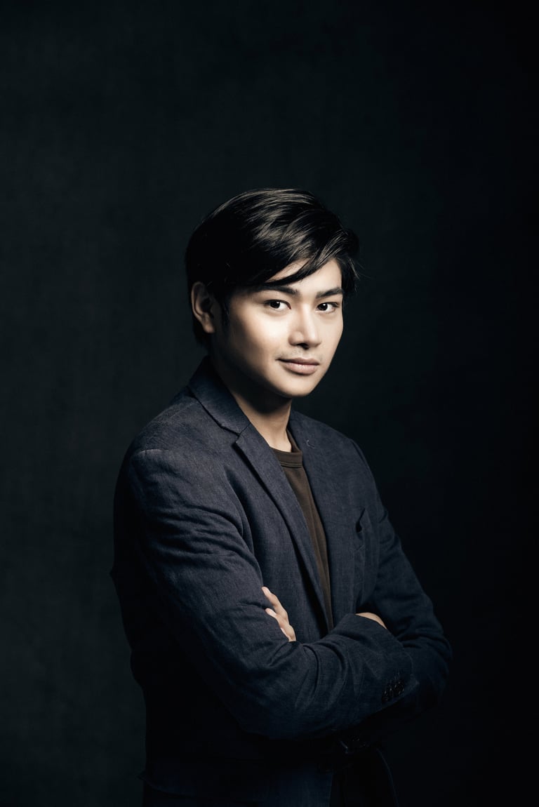 Professional portrait of a young Asian man wearing a dark blazer, arms crossed, against a black background