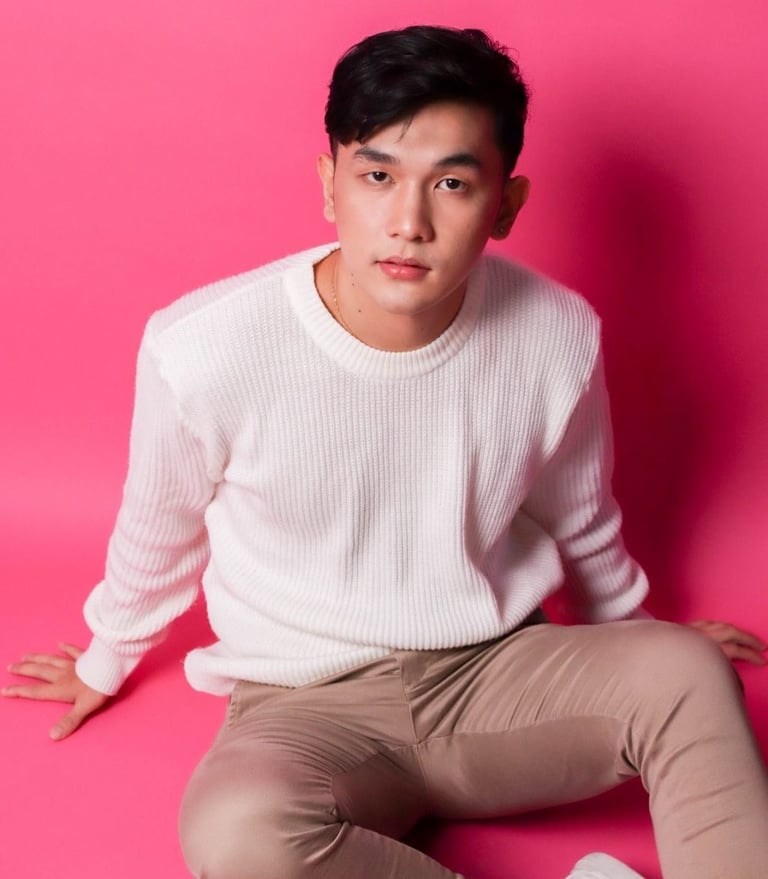 Young man sitting against a bright pink background, wearing a white ribbed sweater and beige pants, looking at camera