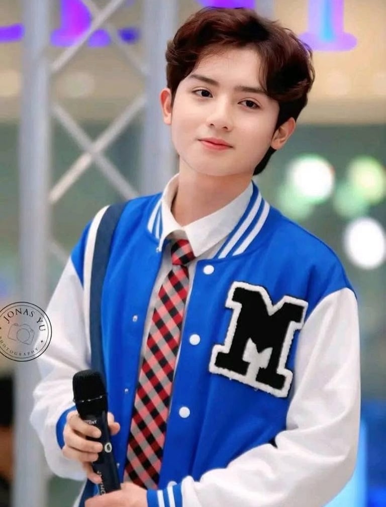 Young boy in blue and white varsity jacket with red plaid tie holding a microphone, smiling at camera