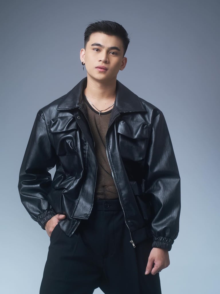 Young man in black leather jacket and mesh shirt posing against gray background