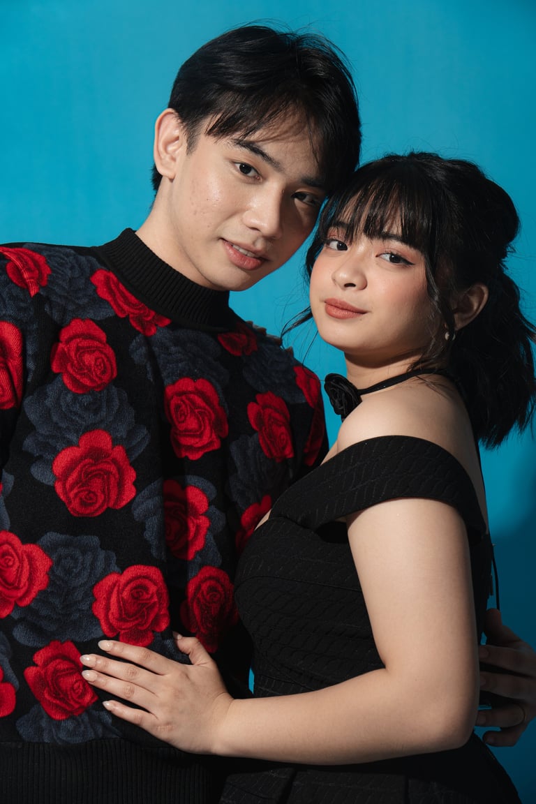 A young man wearing a black sweater with red roses and a young woman in a black off-shoulder top posing together against a bright turquoise background