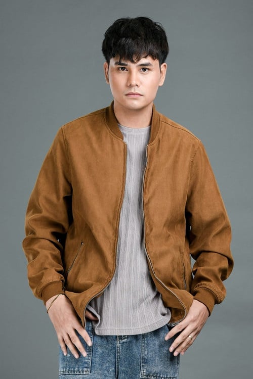 Young man wearing a tan suede jacket over a light striped shirt and blue jeans, posed against a gray background
