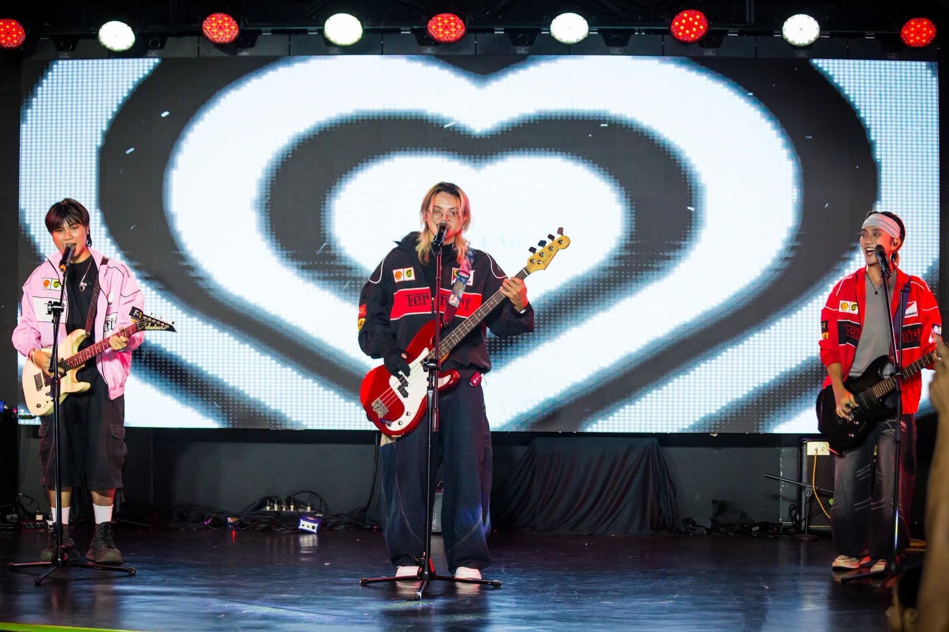 Three musicians performing on stage with guitars in front of a large illuminated heart-shaped screen display