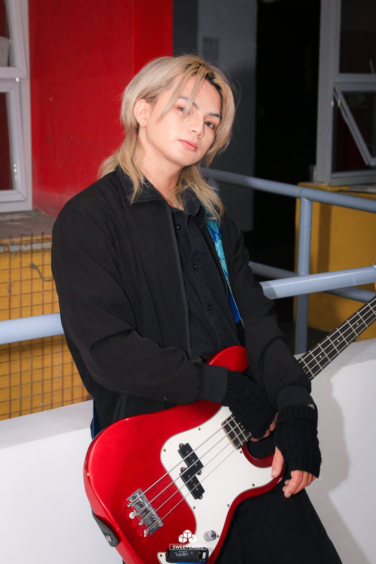 Blonde musician holding a red and white bass guitar, wearing a black jacket and red gloves indoors