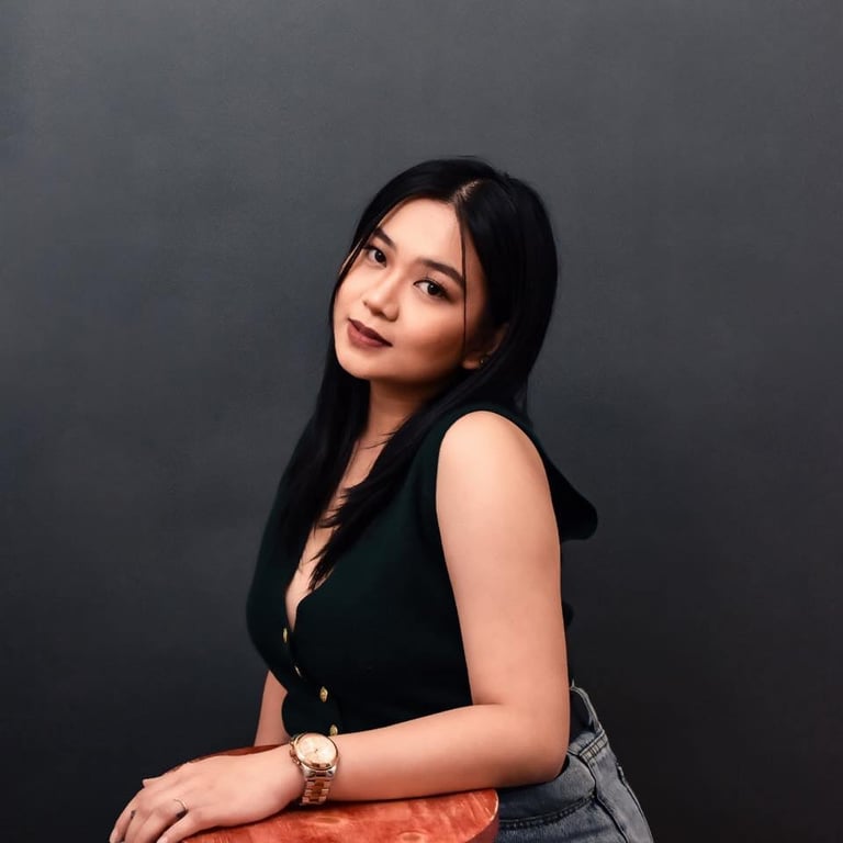 Woman with long black hair wearing a black sleeveless top and denim pants, posing against a gray background