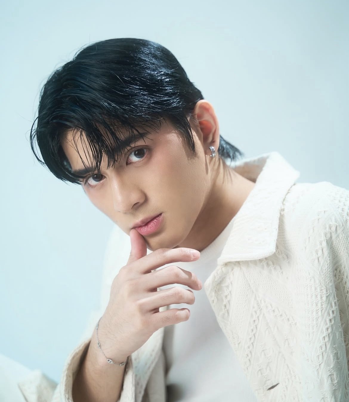 Young man with black hair and pale complexion wearing white textured shirt against light blue background, hand near face in thoughtful pose